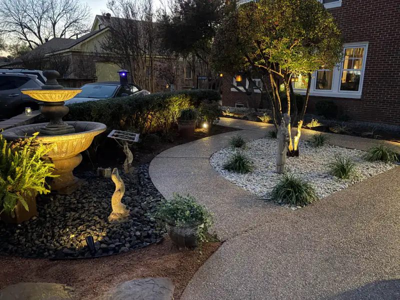 Front landscape at dusk with uplit trees, a tiered fountain and a white rock bed